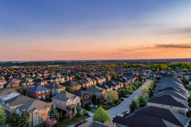 Aerial view of a suburban neighborhood at sunset, featuring rows of houses with rooftops, tree-lined streets, and a colorful sky in the background.