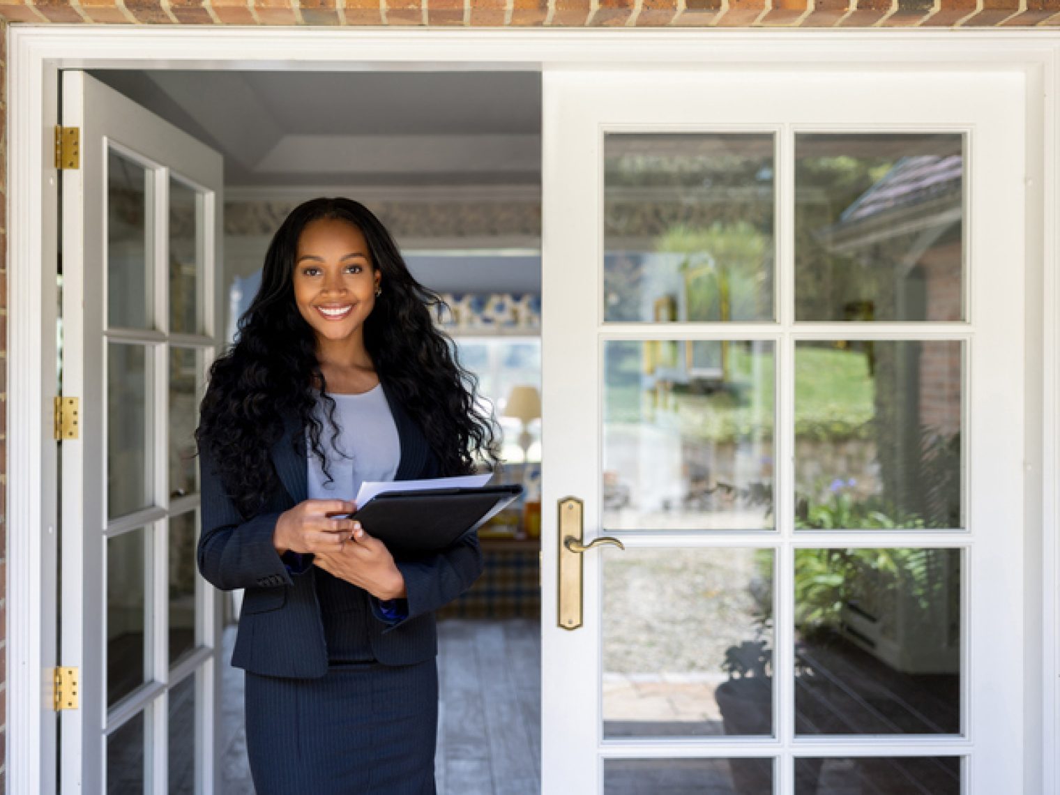 A professional woman with a radiant smile stands at an open door, holding a clipboard, ready to greet or assist, possibly in a real estate context.