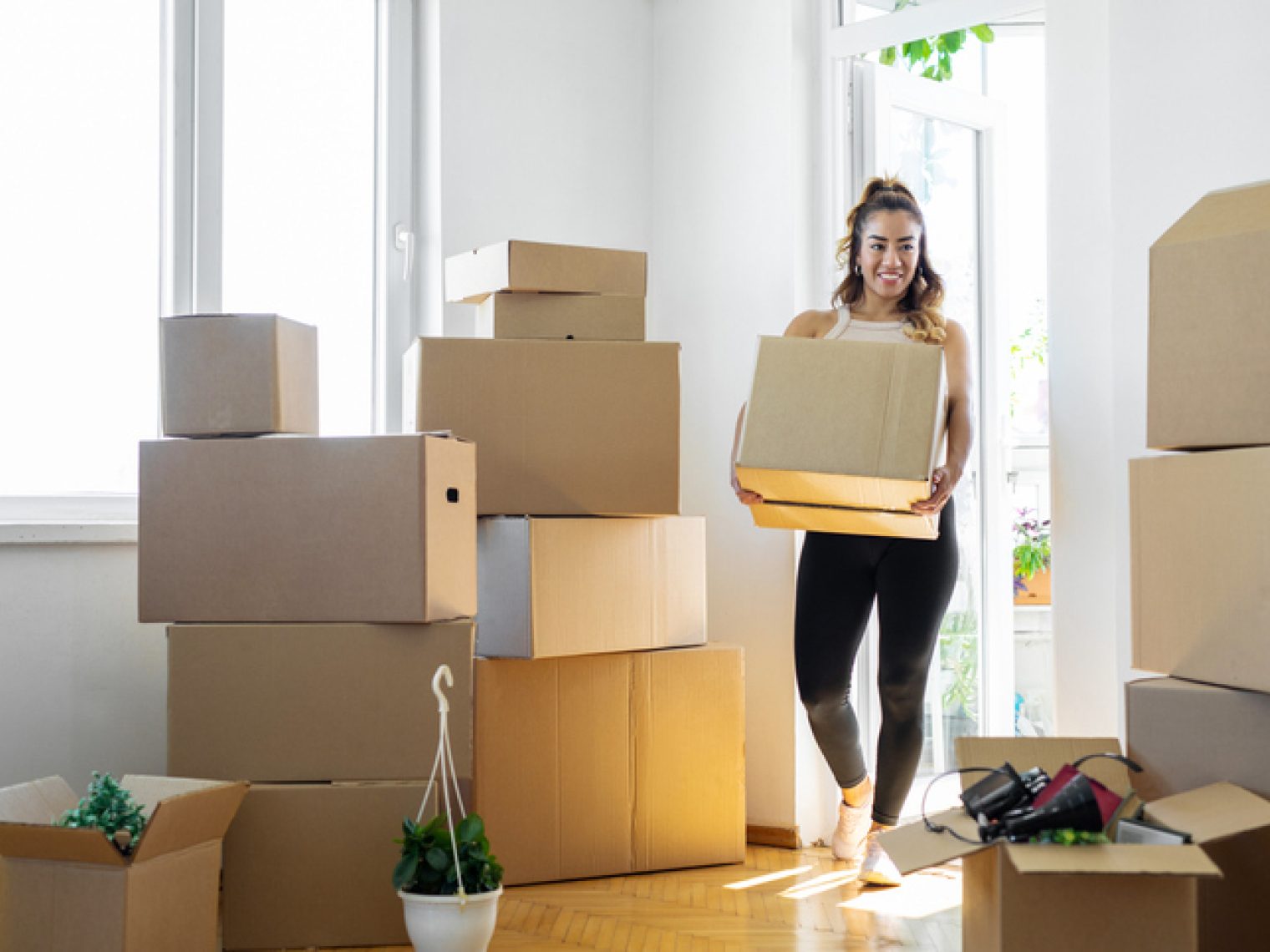 A cheerful woman is carrying a cardboard box in a sunlit room filled with stacked boxes, suggesting a move to a new home. She is wearing a casual outfit suitable for moving day, with a topknot hairstyle, and is smiling, indicating a positive attitude towards the activity.