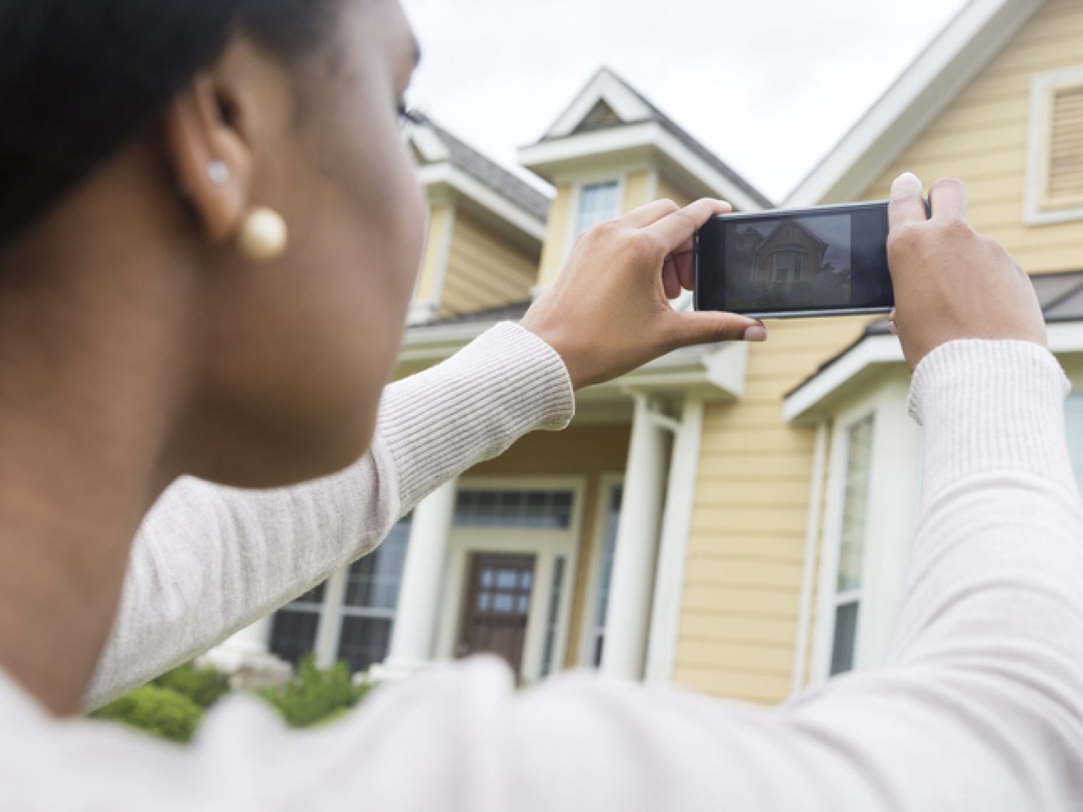 A woman is capturing a photo of a house with her smartphone, potentially for a real estate listing or home inventory.