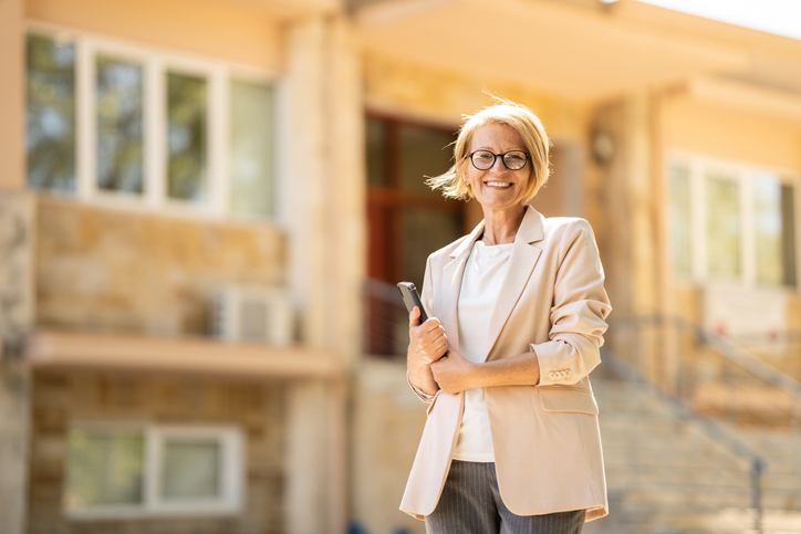 A confident woman in professional attire, holding a tablet, stands smiling in front of a building.