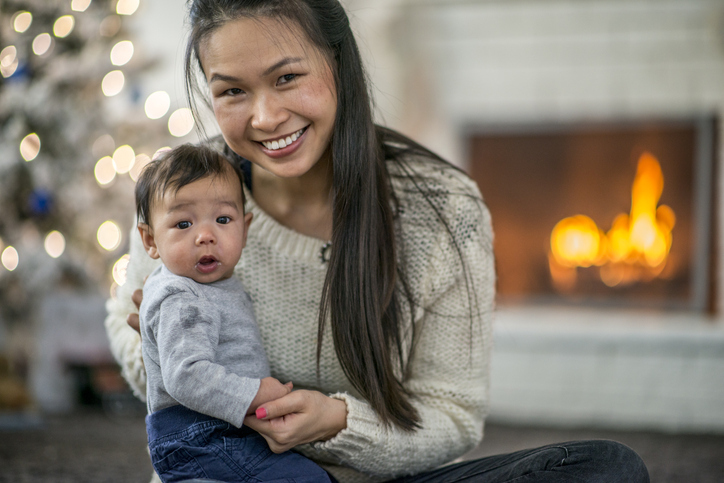 A young woman is holding a baby while sitting in front of a fireplace with a lit fire.