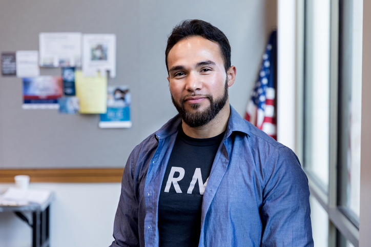 A confident man in a blue shirt over a graphic tee poses for a portrait, with an American flag in the background.